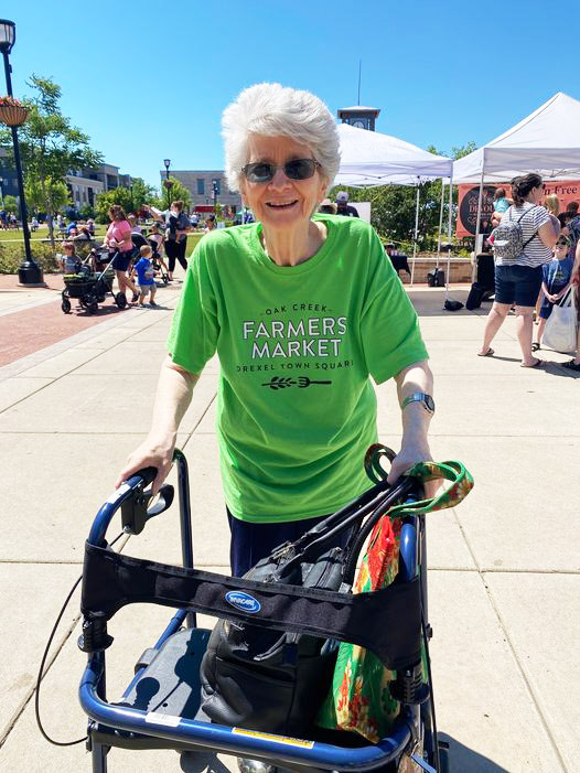 A white-haired woman wearing sunglasses and a green Oak Creek Farmers Market t-shirt smiles broadly as she shops the market with the aid of a blue walker.