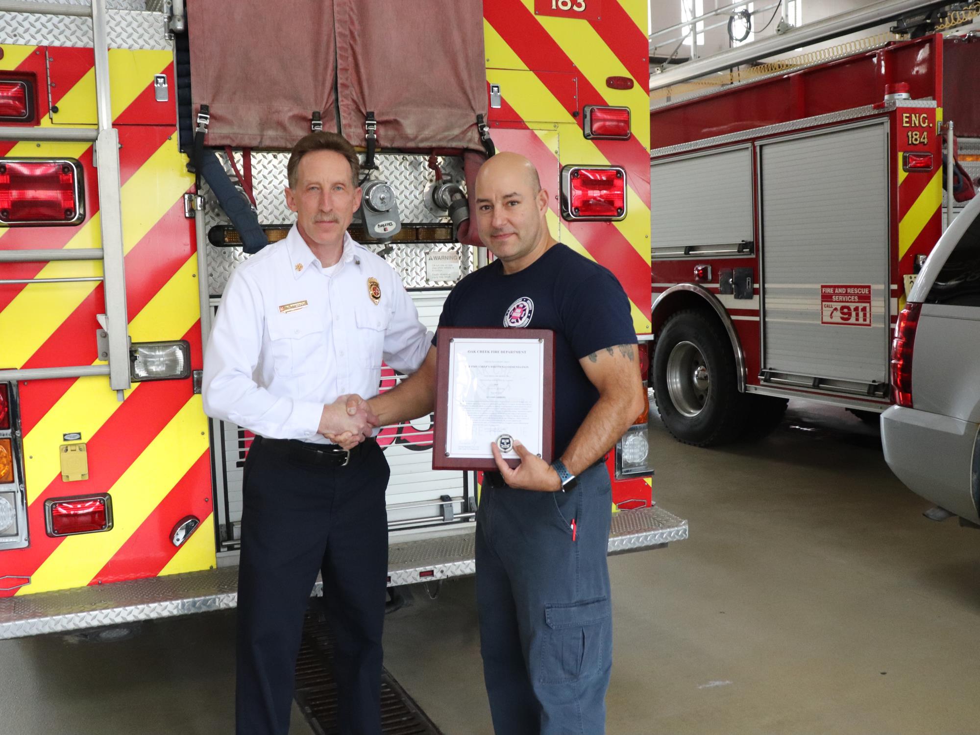 Chief Kressuk and Firefighter Leanna shaking hands while Firefighter Leanna holds Commendation Plaque in front of Engine